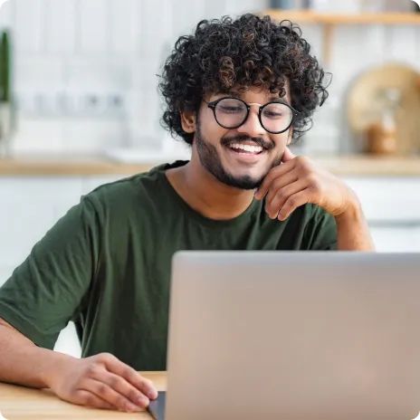 Junger Mann mit lockigem Haar und Brille sitzt lächelnd vor einem Laptop in einer hellen Küche.
