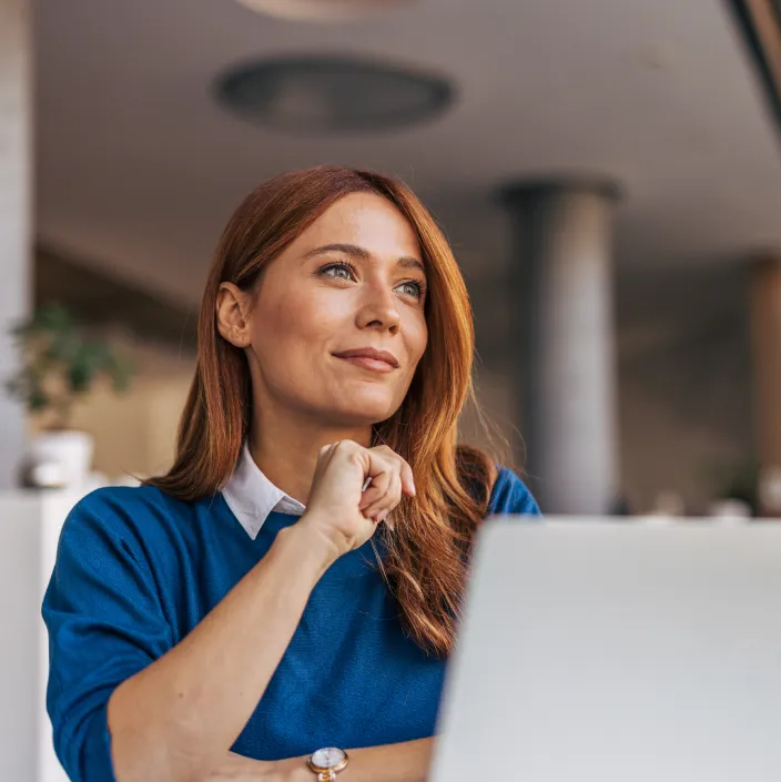 Frau mit roten Haaren sitzt nachdenklich vor einem Laptop in einem modernen Büro und stützt ihr Kinn auf die Hand.