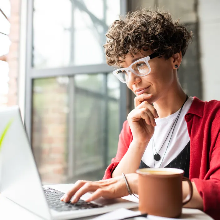 Person mit lockigem kurzem Haar und großen transparenten Brillen arbeitet konzentriert an einem Laptop.