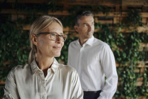 Eine Frau im Vordergrund trägt eine helle Bluse, während ein Mann im Hintergrund steht; beide befinden sich vor einer grünen Wand aus Pflanzen.
