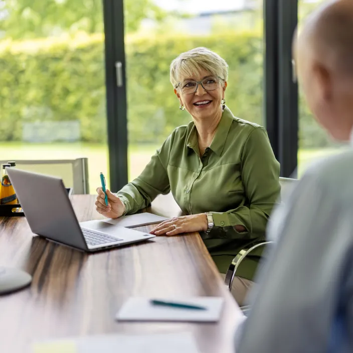Eine Frau mit kurzen grauen Haaren sitzt an einem Holztisch in vor großen Fenstern und grünem Ausblick. Sie trägt ein olivgrünes Hemd und hält einen Stift in der Hand. Vor ihr steht ein Laptop. Im Vordergrund ist unscharf eine zweite Person von hinten zu erkennen.