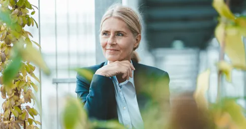Eine Person mit blondem Haar in einem blauen Blazer und Hemd sitzt in einer grünen, lichtdurchfluteten Umgebung. Die Person stützt ihr Kinn auf die Hand, während im Vordergrund unscharfe Pflanzenblätter zu sehen sind. Der Hintergrund zeigt eine moderne Glasstruktur.