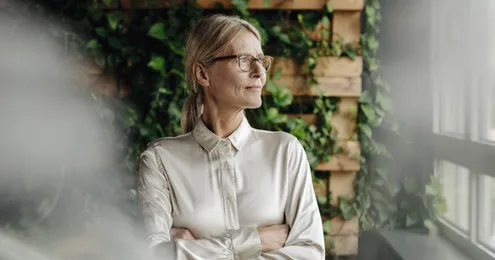 Eine Person in formeller Kleidung steht mit verschränkten Armen vor einer Wand aus Holz und Pflanzen