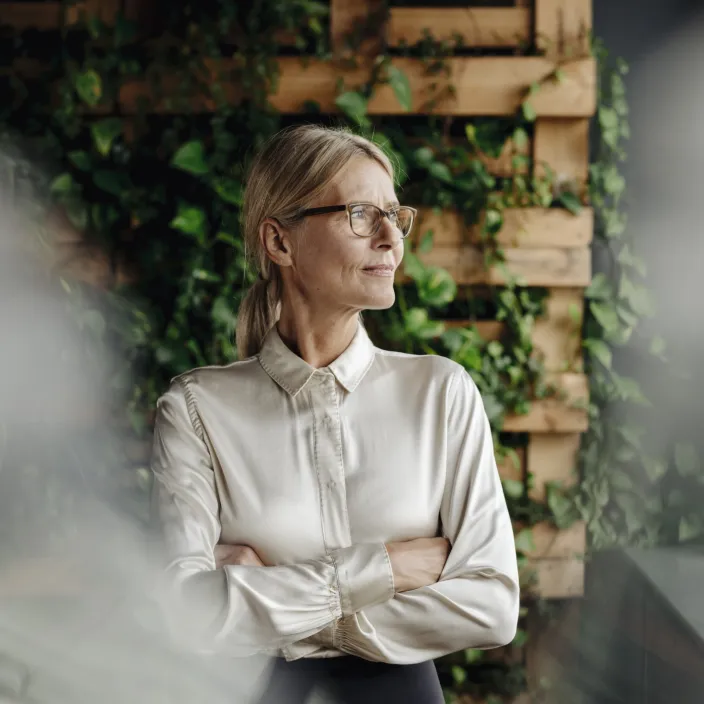 Eine Person in formeller Kleidung steht mit verschränkten Armen vor einer Wand aus Holz und Pflanzen
