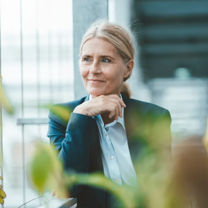 Eine Person mit blondem Haar in einem blauen Blazer und Hemd sitzt in einer grünen, lichtdurchfluteten Umgebung. Die Person stützt ihr Kinn auf die Hand, während im Vordergrund unscharfe Pflanzenblätter zu sehen sind. Der Hintergrund zeigt eine moderne Glasstruktur.