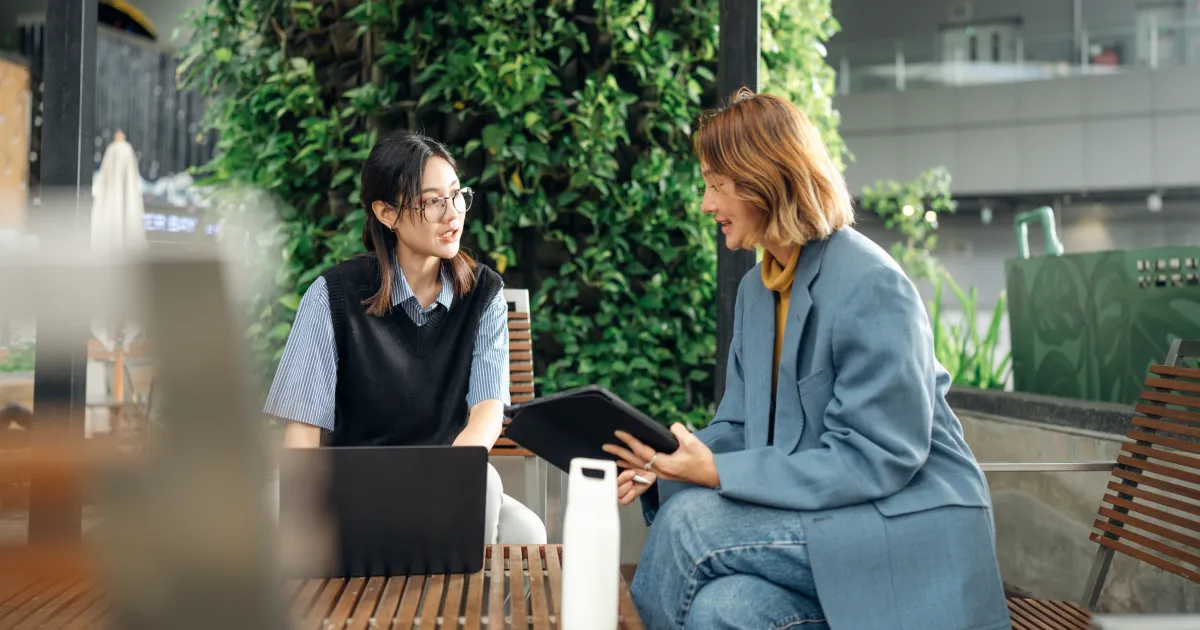 Zwei Personen sitzen auf Holzbänken in einer modernen, begrünten Umgebung. Eine Person hält ein Tablet, während die andere vor einem Laptop sitzt. Im Hintergrund sind Pflanzenwände zu sehen.