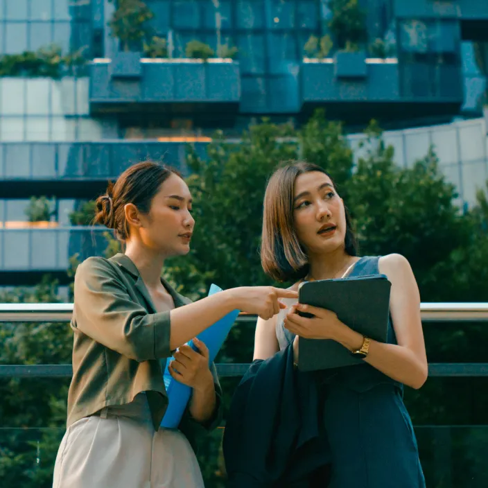 Zwei Kolleginnen besprechen geschäftliche Unterlagen im Freien vor einem modernen Bürogebäude.