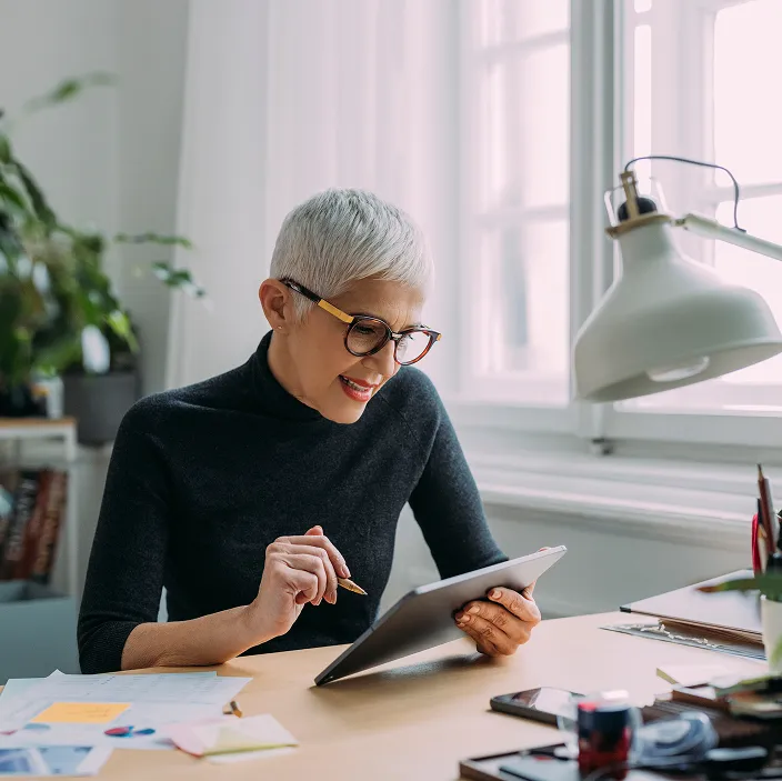 Eine Frau mit kurzen grauen Haaren sitzt an einem Schreibtisch und arbeitet auf einem Tablet. Im Hintergrund ist ein großes Fenster, sowie eine moderne Tischlampe zu sehen.
