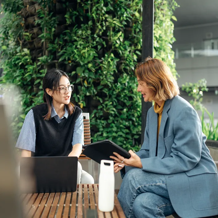 Zwei Personen sitzen auf Holzbänken in einer modernen, begrünten Umgebung. Eine Person hält ein Tablet, während die andere vor einem Laptop sitzt. Im Hintergrund sind Pflanzenwände zu sehen.