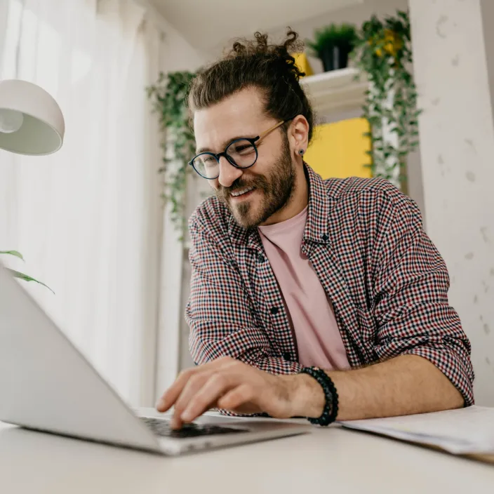 Person mit lockigem Haar und kariertem Hemd arbeitet an einem Laptop an einem Schreibtisch, im Hintergrund Pflanzen und ein Fenster mit hellem Tageslicht.