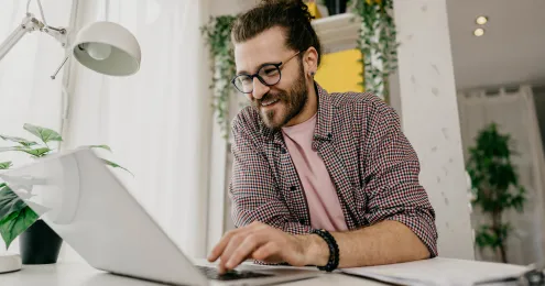 Person mit lockigem Haar und kariertem Hemd arbeitet an einem Laptop an einem Schreibtisch, im Hintergrund Pflanzen und ein Fenster mit hellem Tageslicht.