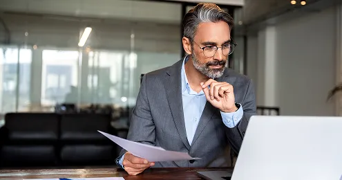 Ein Geschäftsmann in einem grauen Anzug sitzt an einem Schreibtisch mit einem Laptop, hält ein Dokument in der Hand und scheint nachdenklich. Im Hintergrund ist ein modernes Büro zu sehen.