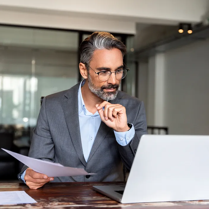 Ein Geschäftsmann in einem grauen Anzug sitzt an einem Schreibtisch mit einem Laptop, hält ein Dokument in der Hand und scheint nachdenklich. Im Hintergrund ist ein modernes Büro zu sehen.