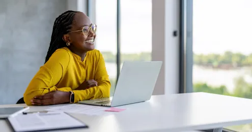 Eine Person mit geflochtenem Haar sitzt an einem Schreibtisch vor einem Laptop. Sie trägt ein senfgelbes Oberteil und eine Smartwatch.