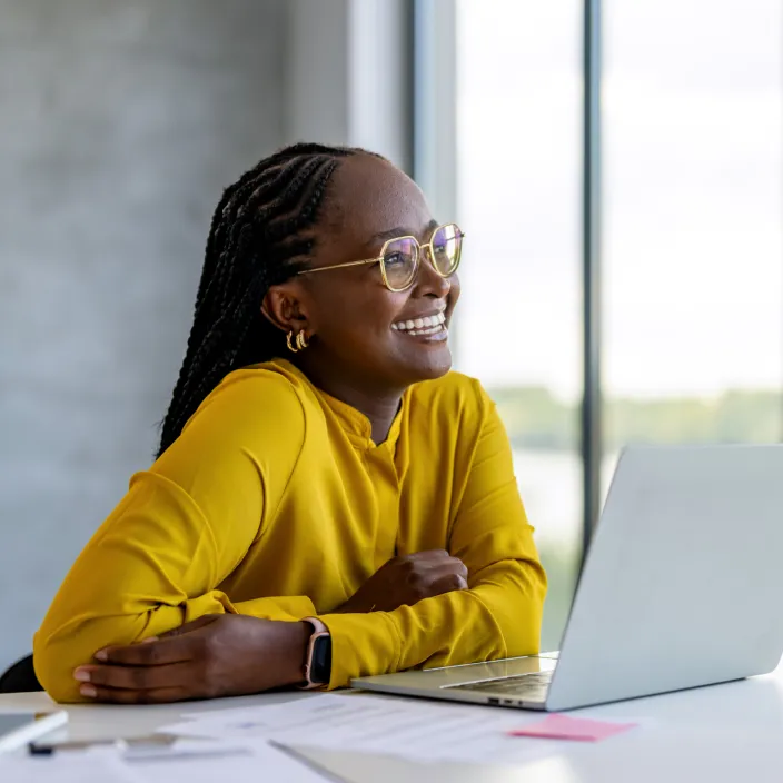 Eine Person mit geflochtenem Haar sitzt an einem Schreibtisch vor einem Laptop. Sie trägt ein senfgelbes Oberteil und eine Smartwatch.