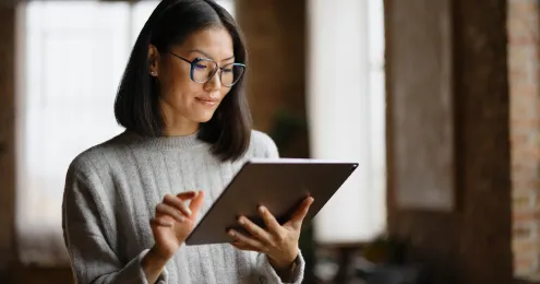 Eine Person mit schulterlangem, glattem, dunklem Haar trägt einen grauen Pullover und hält ein Tablet in den Händen. Die Umgebung ist warm beleuchtet mit einem Hintergrund, der auf einen Innenraum hindeutet.