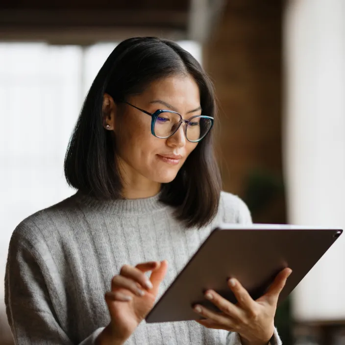Eine Person mit schulterlangem, glattem, dunklem Haar trägt einen grauen Pullover und hält ein Tablet in den Händen. Die Umgebung ist warm beleuchtet mit einem Hintergrund, der auf einen Innenraum hindeutet.