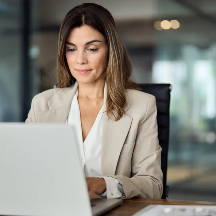 Eine Frau mit offenem Haar sitzt an einem Schreibtisch vor einem Laptop. Sie trägt einen beigen Blazer über einer weißen Bluse.