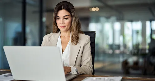 Eine Frau mit offenem Haar sitzt an einem Schreibtisch vor einem Laptop. Sie trägt einen beigen Blazer über einer weißen Bluse.