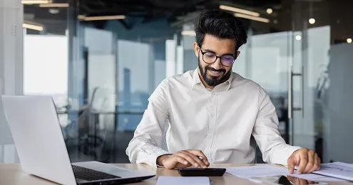 Eine Person in einem weißen Hemd sitzt an einem Schreibtisch mit einem Laptop, Smartphone und Dokumenten. Im Hintergrund ist ein modernes Büro mit Glaswänden sichtbar.