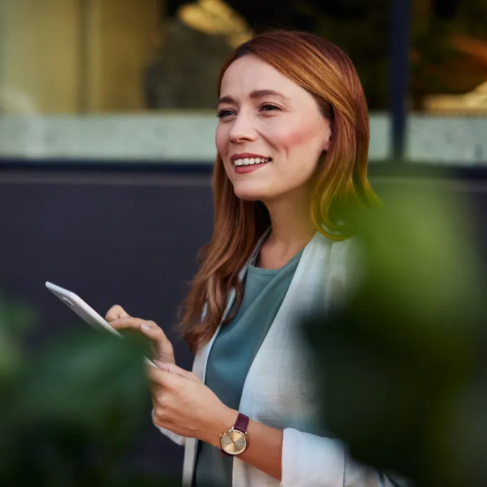 Eine rothaarige Frau mittleren Alters, gekleidet in eine hellgraue Jacke und ein grünes Oberteil, die im Freien eine weiße Tablette oder ein Smartphone hält und nach rechts oben blickt
