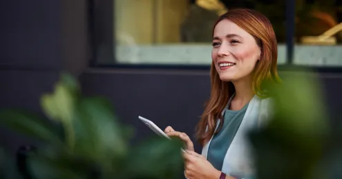 Eine rothaarige Frau mittleren Alters, gekleidet in eine hellgraue Jacke und ein grünes Oberteil, die im Freien eine weiße Tablette oder ein Smartphone hält und nach rechts oben blickt