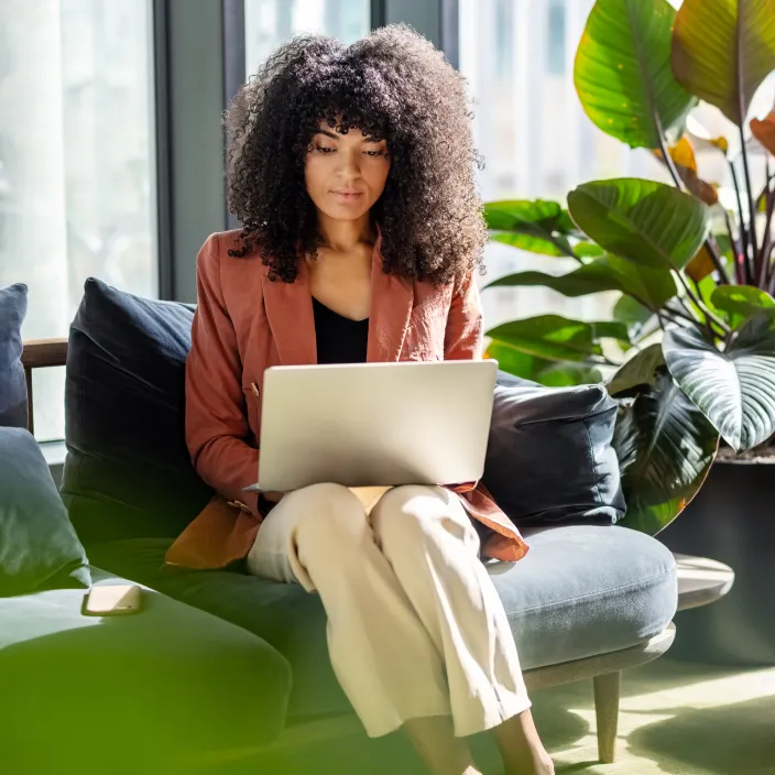 Eine Person sitzt in einem modernen, lichtdurchfluteten Raum auf einem Sofa und arbeitet an einem Laptop. Sie trägt einen rostfarbenen Blazer und helle Hosen.