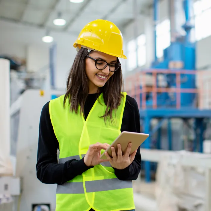 Eine Person in einem industriellen Umfeld trägt eine gelbe Sicherheitsweste und einen gelben Schutzhelm. Sie hält ein Tablet in den Händen.