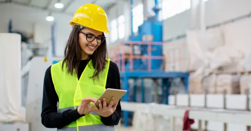 Eine Person in einem industriellen Umfeld trägt eine gelbe Sicherheitsweste und einen gelben Schutzhelm. Sie hält ein Tablet in den Händen.