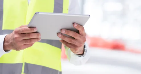 Ein Mann in einem hellblauen Hemd steht in einer modernen Fabrikumgebung und hält ein Tablet in der Hand.