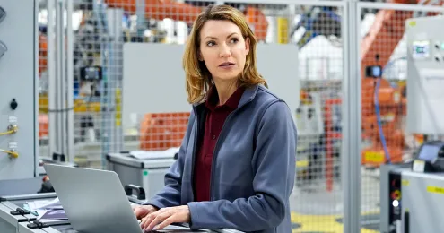 Eine Frau steht in einer modernen Fabrikhalle an einem mobilen Arbeitsplatz und bedient einen Laptop.