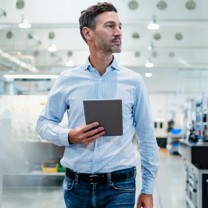 Ein Mann in einem hellblauen Hemd steht in einer modernen Fabrikumgebung und hält ein Tablet in der Hand.