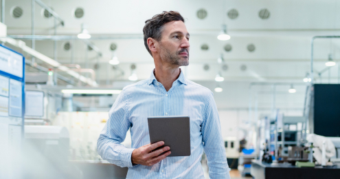 Ein Mann in einem hellblauen Hemd steht in einer modernen Fabrikumgebung und hält ein Tablet in der Hand.
