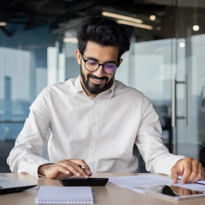 Eine Person in einem weißen Hemd sitzt an einem Schreibtisch mit einem Laptop, Smartphone und Dokumenten. Im Hintergrund ist ein modernes Büro mit Glaswänden sichtbar.