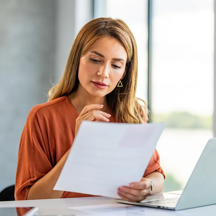 Eine Person in einem orangefarbenen Oberteil sitzt an einem Schreibtisch, hält ein Dokument in der Hand und arbeitet an einem Laptop. Im Hintergrund sind große Fenster mit Blick auf eine helle, grüne Landschaft zu sehen.