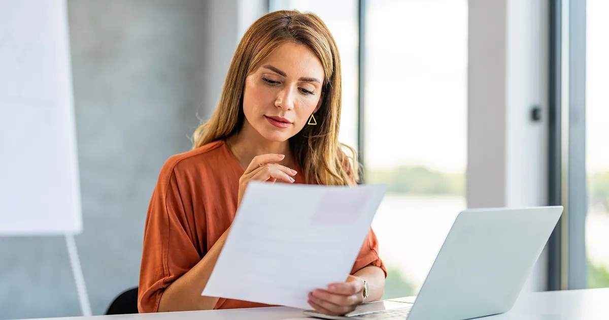 Eine Person in einem orangefarbenen Oberteil sitzt an einem Schreibtisch, hält ein Dokument in der Hand und arbeitet an einem Laptop. Im Hintergrund sind große Fenster mit Blick auf eine helle, grüne Landschaft zu sehen.