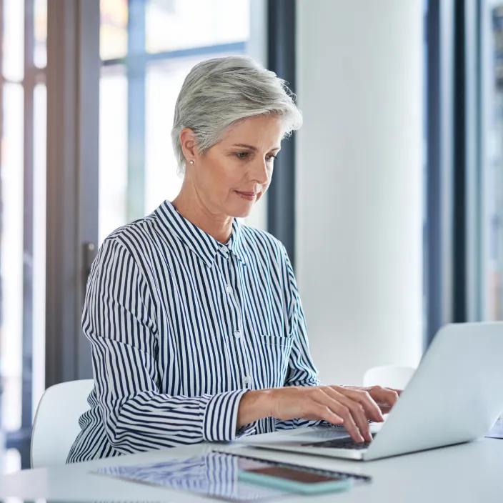 Eine Person mit grauem Haar und gestreifter Bluse arbeitet an einem Laptop in einem modernen, lichtdurchfluteten Büro. Auf dem Tisch liegen Dokumente, ein Smartphone und eine Brille.