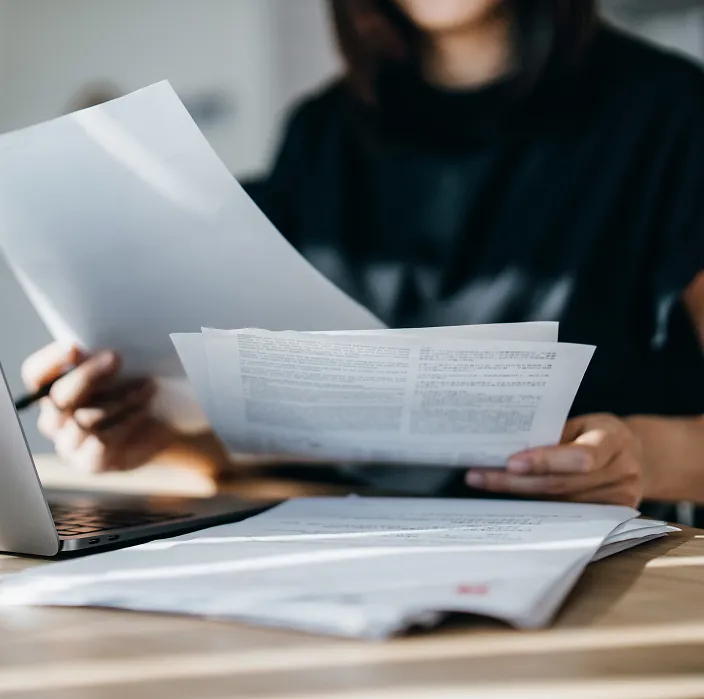 Eine Person sitzt an einem Schreibtisch und hält mehrere Dokumente in den Händen. Auf dem Tisch liegen weitere Papiere sowie ein aufgeklappter Laptop.