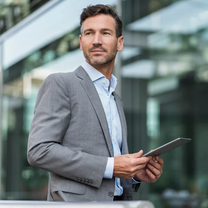 Ein Mann in einem grauen Anzug und hellblauem Hemd hält ein Tablet in den Händen. Er steht vor einer modernen Glasfassade, die einen unscharfen Hintergrund mit Reflexionen zeigt.