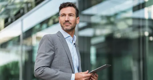 Ein Mann in einem grauen Anzug und hellblauem Hemd hält ein Tablet in den Händen. Er steht vor einer modernen Glasfassade, die einen unscharfen Hintergrund mit Reflexionen zeigt.