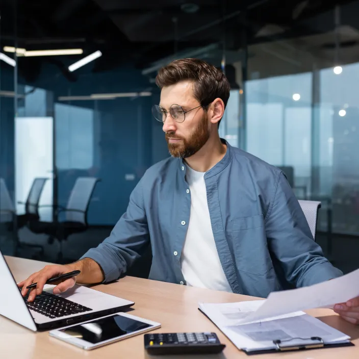 Ein Mann sitzt an einem Schreibtisch in einem modernen Büro mit Glaswänden. Er trägt ein hellblaues Hemd über einem weißen T-Shirt und arbeitet am Laptop, während er ein Dokument in der Hand hält.
