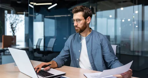 Ein Mann sitzt an einem Schreibtisch in einem modernen Büro mit Glaswänden. Er trägt ein hellblaues Hemd über einem weißen T-Shirt und arbeitet am Laptop, während er ein Dokument in der Hand hält.
