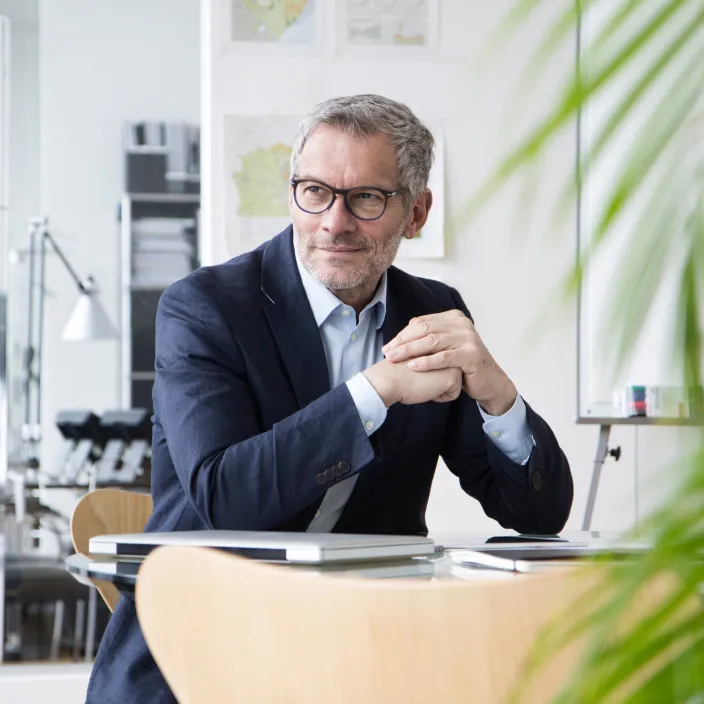 Ein Mann mit Brille und kurzem, grauem Haar sitzt in einem hellen, modernen Büro an einem Tisch.