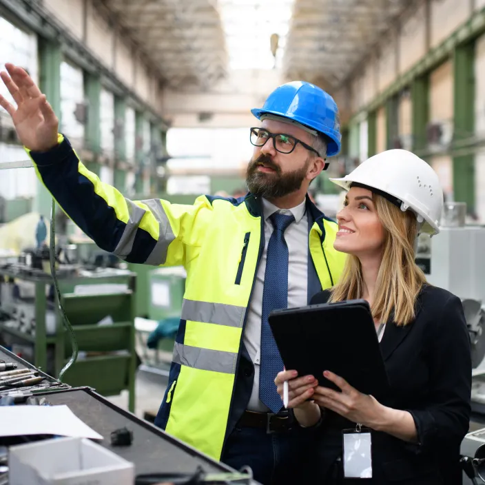 Ein Ingenieur in einer gelben Warnjacke und blauem Schutzhelm zeigt auf etwas in einer großen Fabrikhalle, während er einer Frau mit weißem Schutzhelm und einem Tablet in der Hand etwas erklärt.