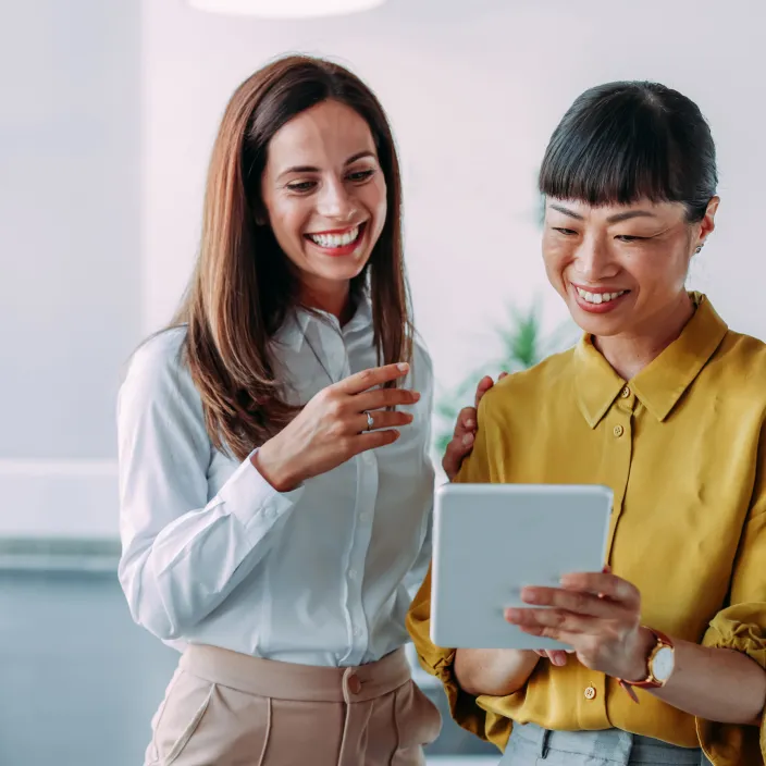Zwei lächelnde Frauen in einem Büro schauen gemeinsam auf ein Tablet. Eine Frau trägt eine hellblaue Bluse, die andere eine goldgelbe Bluse und eine Armbanduhr.