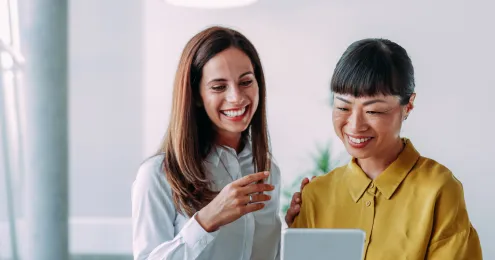 Zwei lächelnde Frauen in einem Büro schauen gemeinsam auf ein Tablet. Eine Frau trägt eine hellblaue Bluse, die andere eine goldgelbe Bluse und eine Armbanduhr.