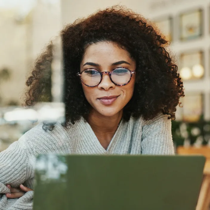 Eine junge Frau mit lockigem Haar und runder Brille schaut konzentriert auf einen Laptop. Sie trägt einen hellgrauen Strickpullover und sitzt in einem hell eingerichteten Café oder Büro.