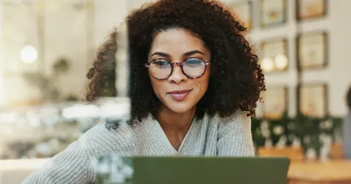 Eine junge Frau mit lockigem Haar und runder Brille schaut konzentriert auf einen Laptop. Sie trägt einen hellgrauen Strickpullover und sitzt in einem hell eingerichteten Café oder Büro.