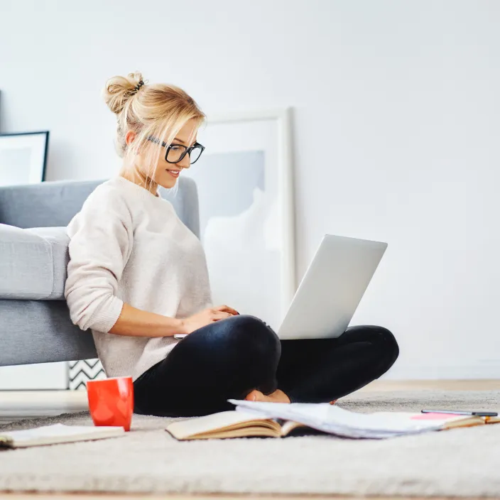 Eine junge blonde Frau mit hochgestecktem Haar und schwarzer Brille sitzt mit gekreuzten Beinen auf einem hellen Teppich, angelehnt an ein graues Sofa. Sie arbeitet an einem Laptop.