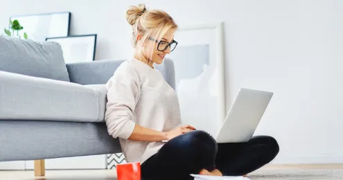 Eine junge blonde Frau mit hochgestecktem Haar und schwarzer Brille sitzt mit gekreuzten Beinen auf einem hellen Teppich, angelehnt an ein graues Sofa. Sie arbeitet an einem Laptop.